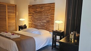 A neatly made bed with white linens and a brown throw blanket, flanked by two bedside tables with lamps. A wooden wardrobe with horizontal slats is to the left, and a wooden wall paneling is behind the bed. - Bungalow F2 deluxe in beautiful Art Deco property and tropical garden (Petit Bourg)