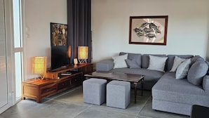 A modern living room with a grey sofa, ottomans, a wooden coffee table, a TV, and a floor lamp. - Bungalow F2 deluxe in beautiful Art Deco property and tropical garden (Petit Bourg)