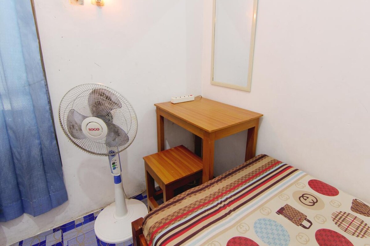 A small room with a bed, a wooden table, a standing fan, and a mirror on the wall.