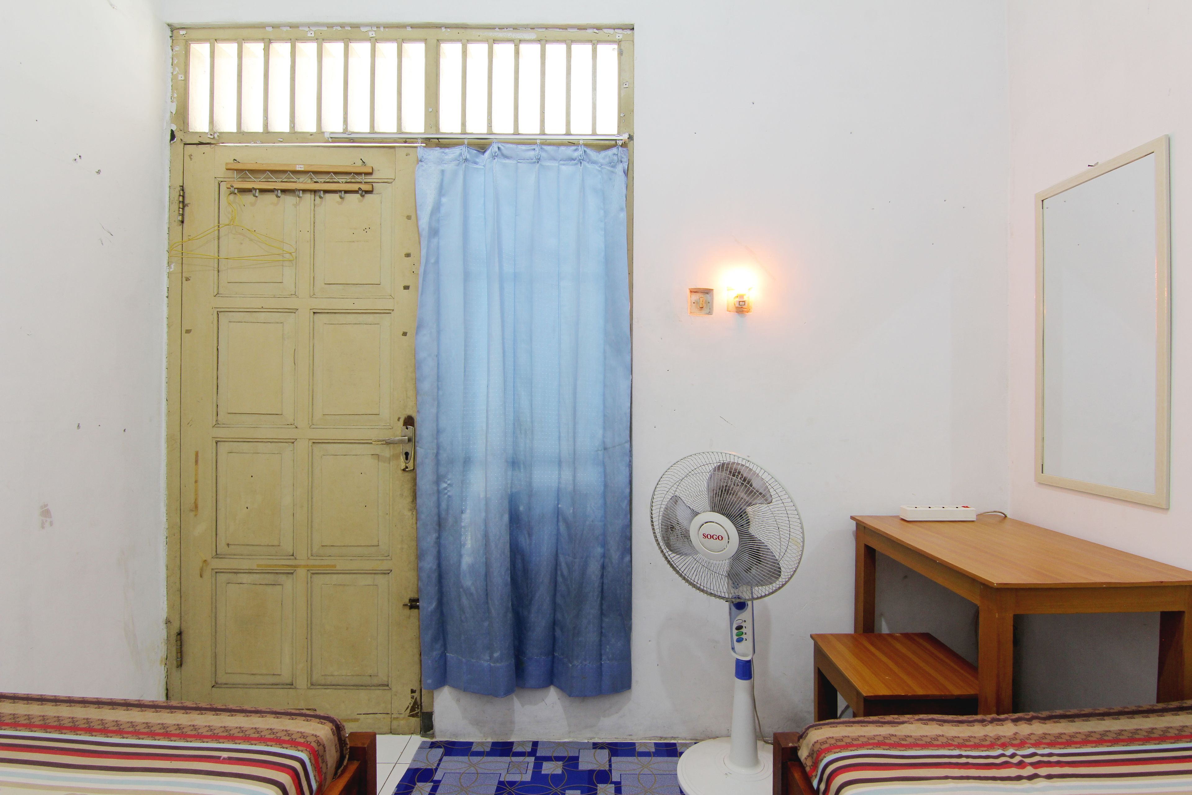 Una habitación con una cama, una mesa de madera, un ventilador de pie, una lámpara y una puerta con cortina.