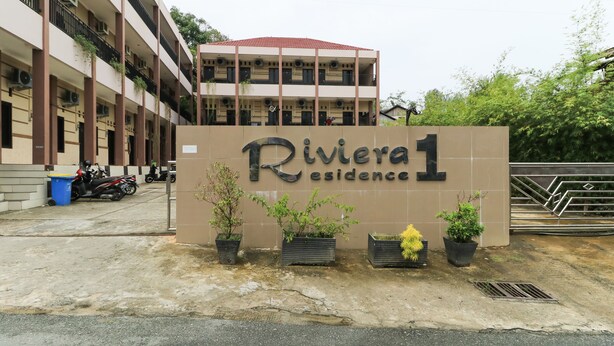 A building with a sign reading "Riviera 1 Residence" and several potted plants in front.