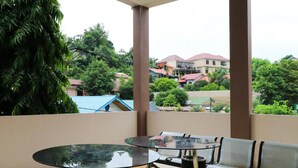 A balcony with a glass table and chairs, a trash bin, and a view of residential buildings and greenery.