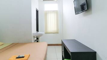 Een kleine, schone kamer met een bed, een aan de muur bevestigde televisie, een raam met gordijnen, een wastafel en een groene prullenbak.