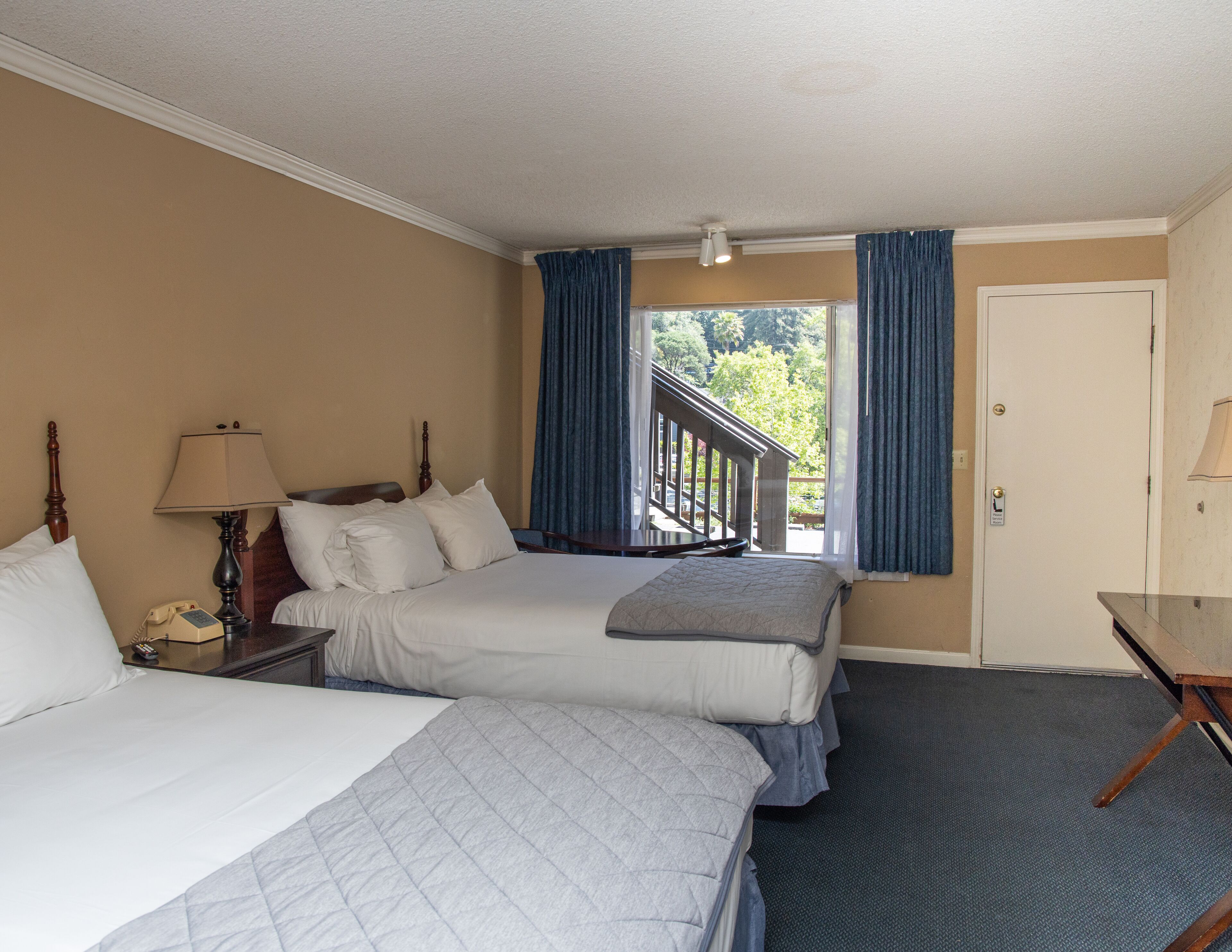 A hotel room with two beds, a desk, a lamp, and a view of the outdoors.