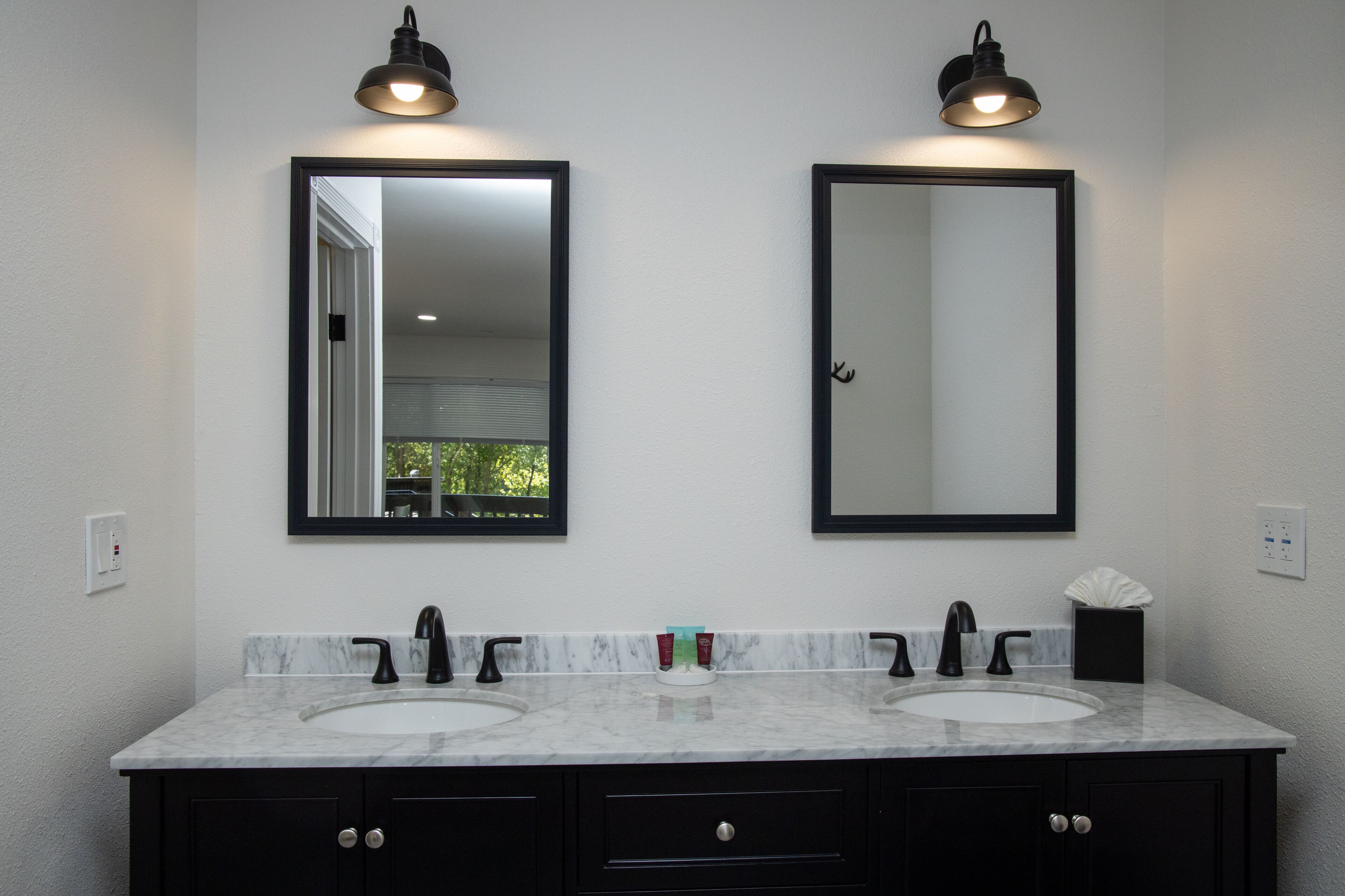 Une salle de bain avec deux vasques, des robinets noirs et un plan de toilette en marbre. Deux miroirs muraux, dotés de cadres noirs et d’un éclairage situé au-dessus, y sont installés.