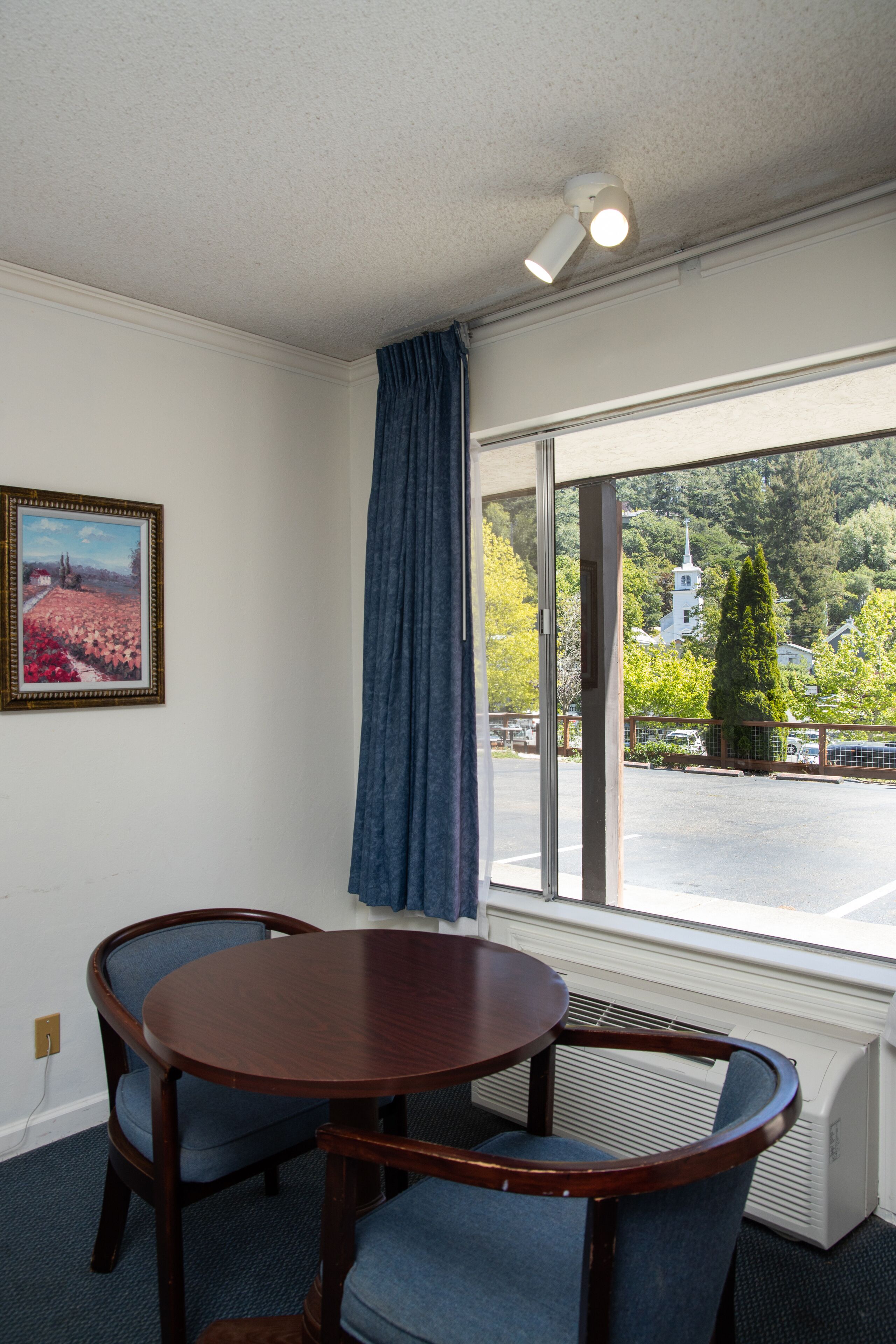 Une chambre d’hôtel avec une table et des chaises, une fenêtre donnant sur un parking et des arbres, et un tableau encadré au mur.