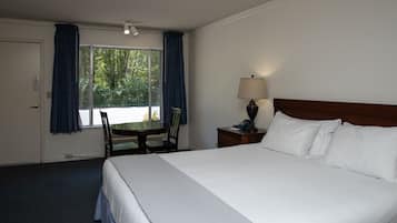 A hotel room with a large bed, a desk with a chair, a lamp, and a view of greenery through the window.