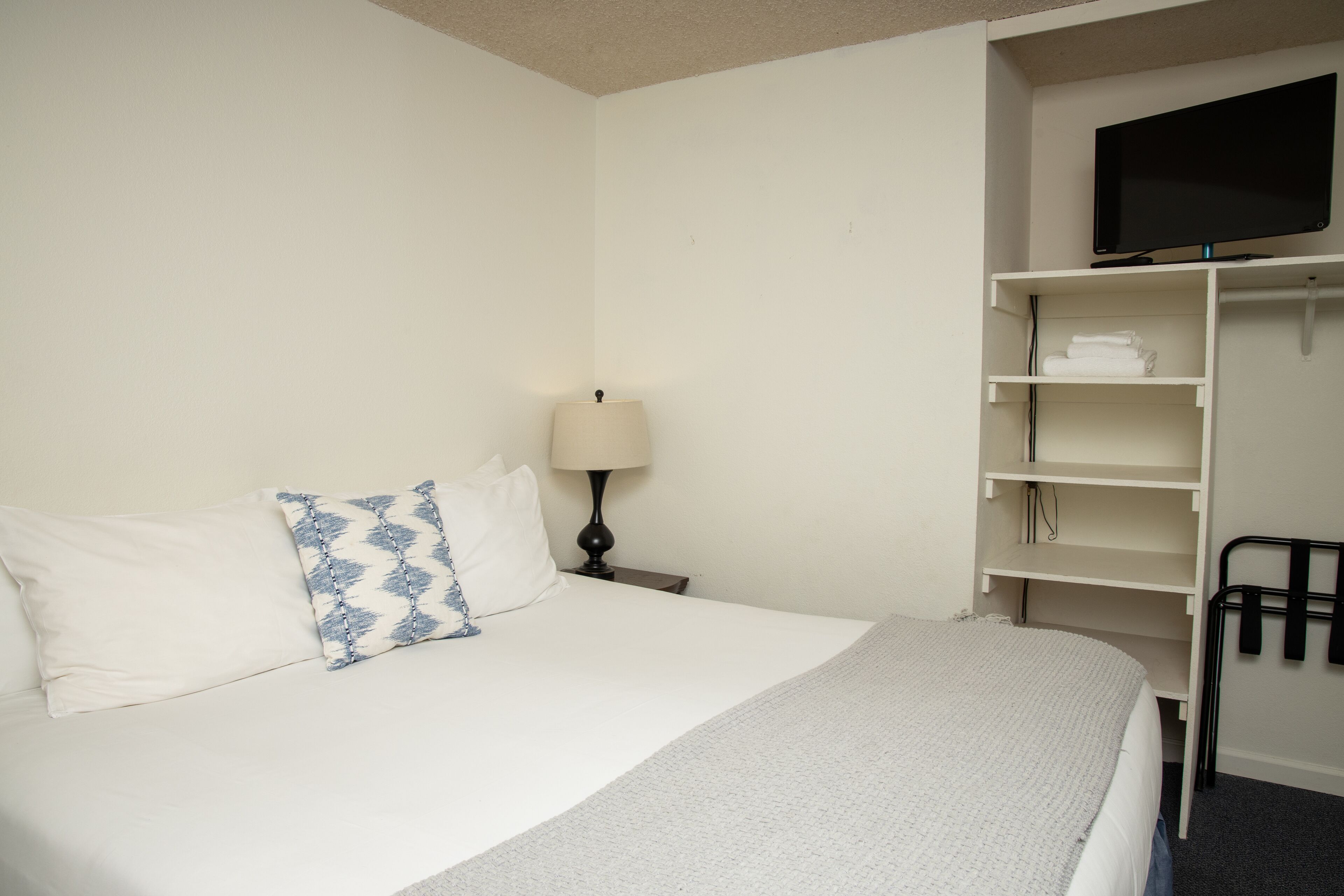 Une chambre d’hôtel avec un lit, un téléviseur posé sur un meuble, une lampe et une armoire avec des étagères.