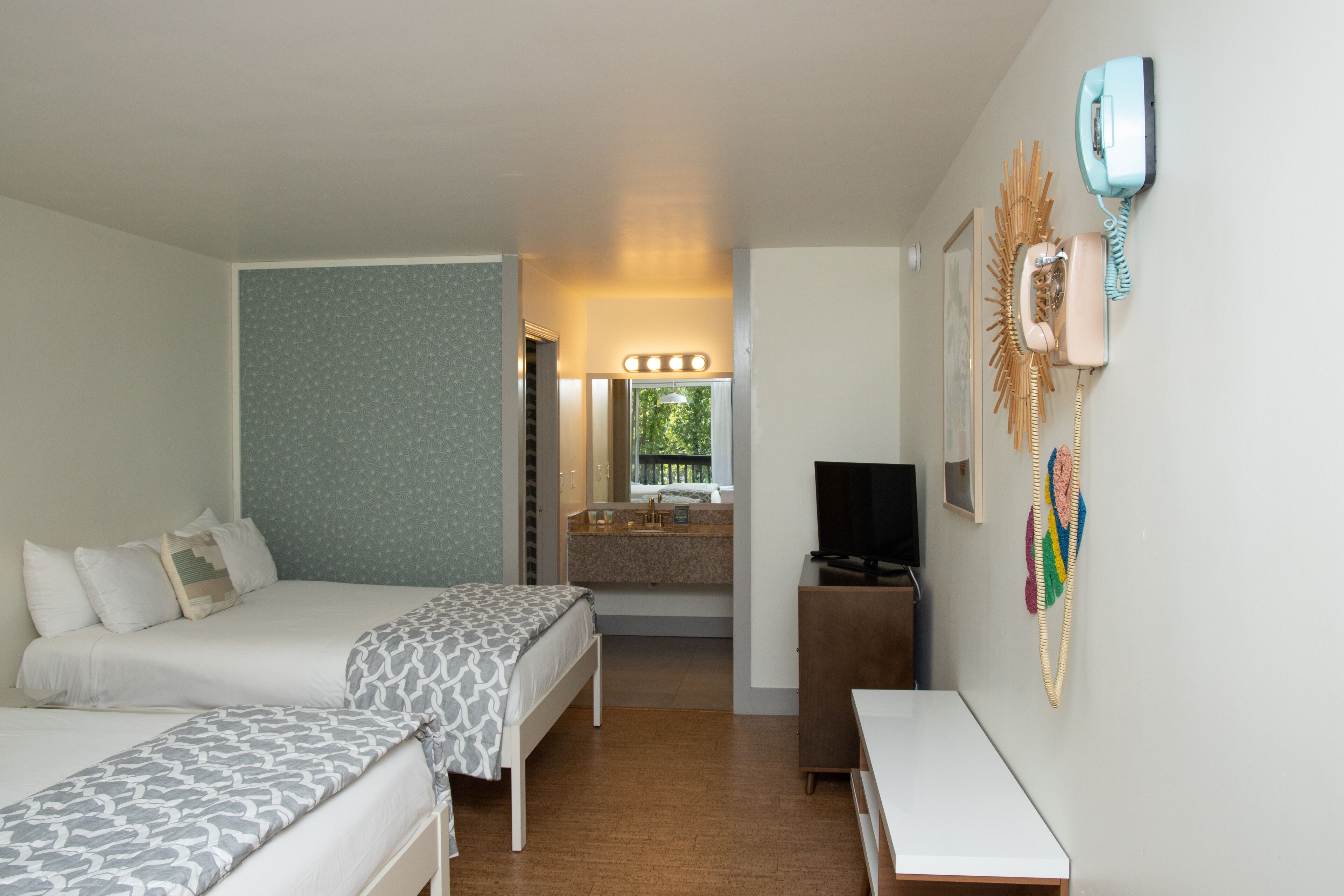 A hotel room with two beds, a TV, a desk, a bathroom with a sink and mirror, and a phone on the wall.