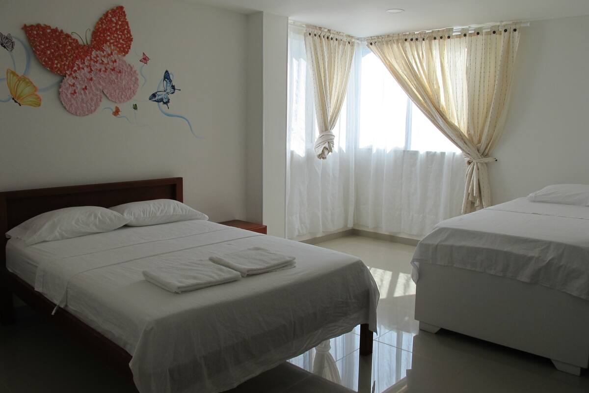 A hotel room with two beds, a large butterfly artwork on the wall, and sheer curtains on the windows.