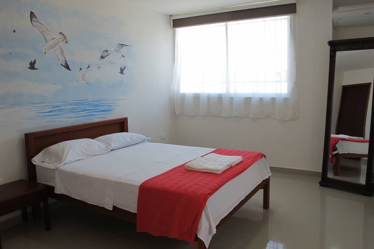 A hotel room with a bed, a window with curtains, a wall mural, and a mirror.