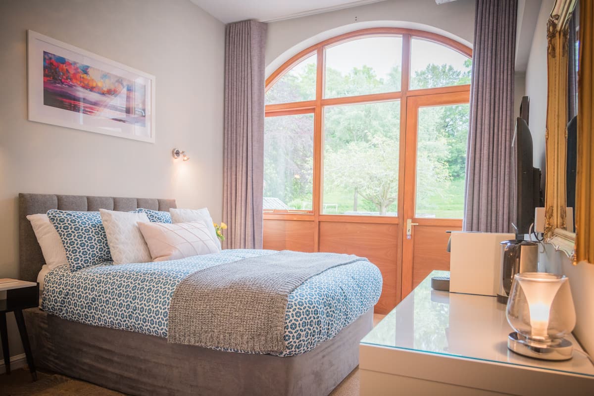 A bedroom with a large bed, a desk with a lamp, a painting on the wall, and a window with curtains.