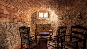 Una habitación con paredes de piedra, una mesa redonda y cuatro sillas, una ventana pequeña con cortinas y cuadros en las paredes.