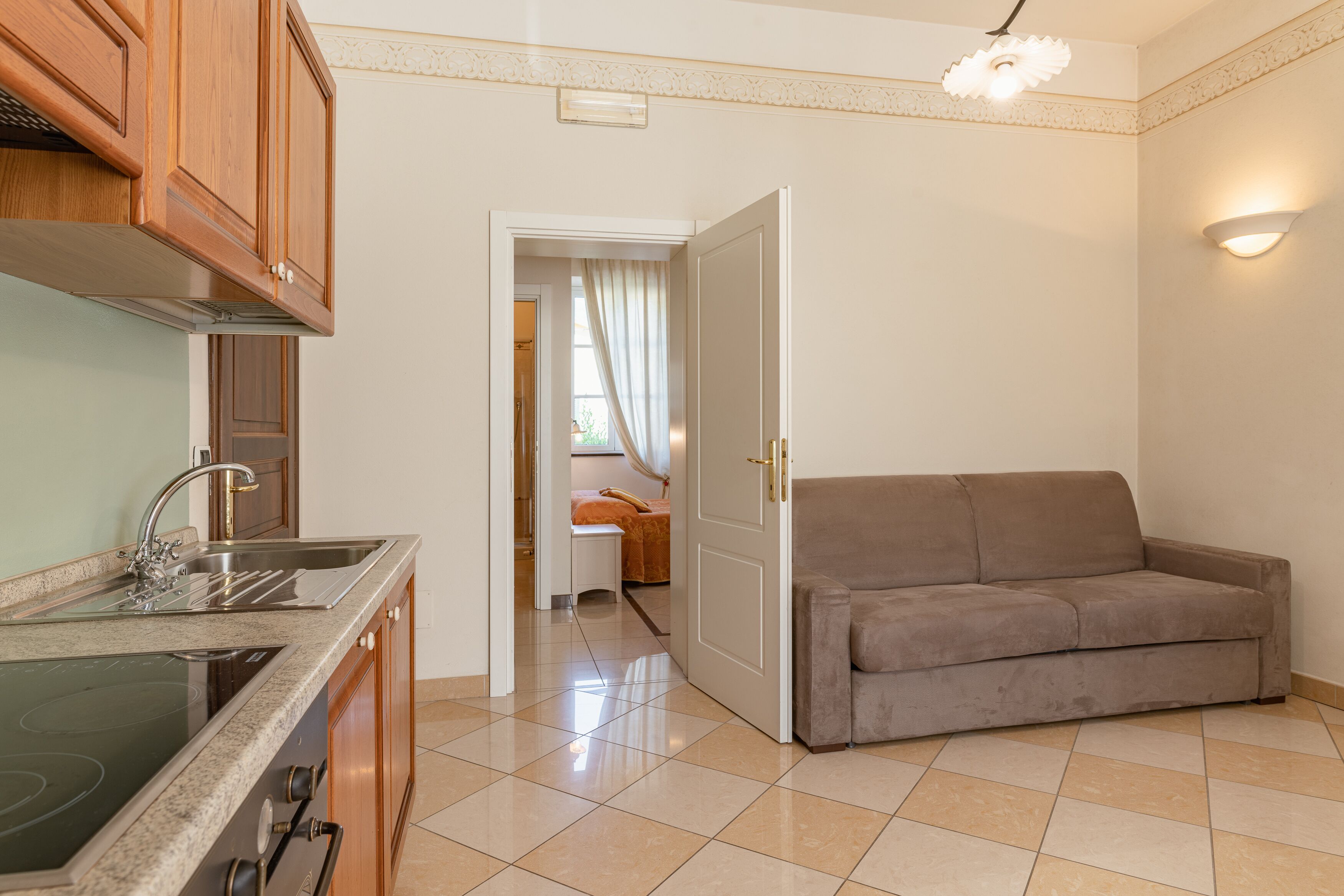 A kitchen with a stove, sink, and wooden cabinets. There is a beige couch and a door leading to a bedroom.