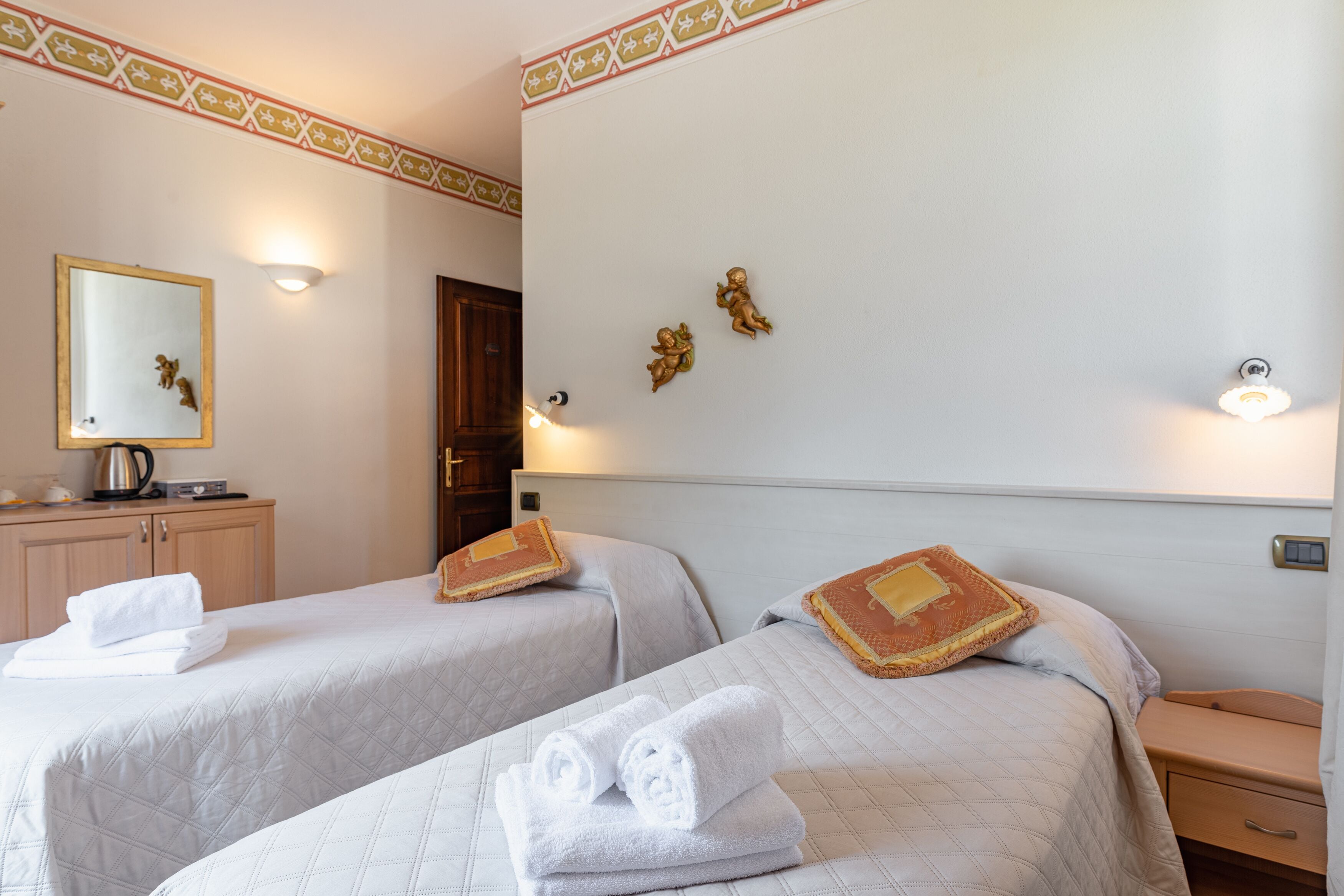 A hotel room with two beds, each with a neatly folded towel, a small bedside table, and a nightstand. There is a mirror on the wall, a decorative wall piece, and a light fixture above the beds.