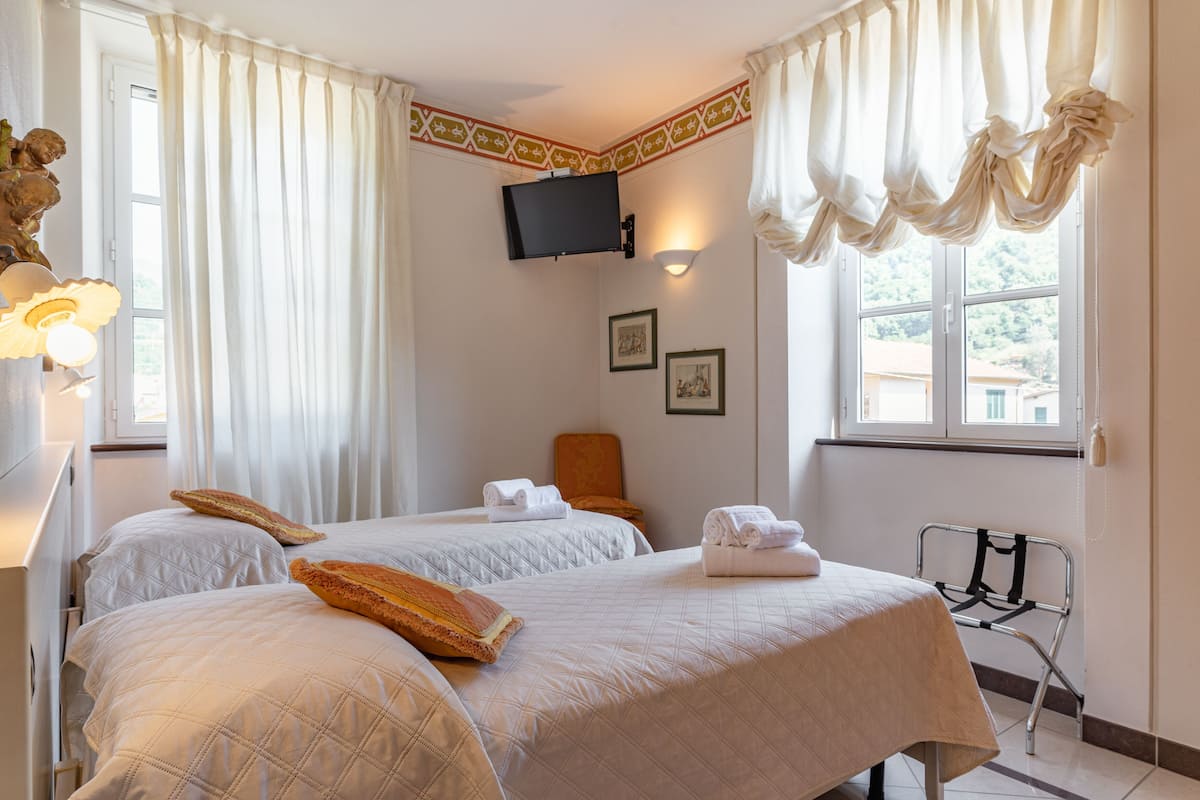 Habitación de hotel con dos camas, un televisor de pantalla plana montado en la pared, una silla y un ventanal con cortinas.