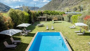 Una piscina con sillas de descanso y sombrillas, rodeada de un césped bien cuidado y un pabellón de madera.