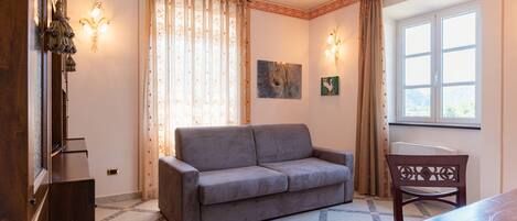 A room with a grey sofa, a wooden chair, a dining table, a window with curtains, wall-mounted lights, and a painting.
