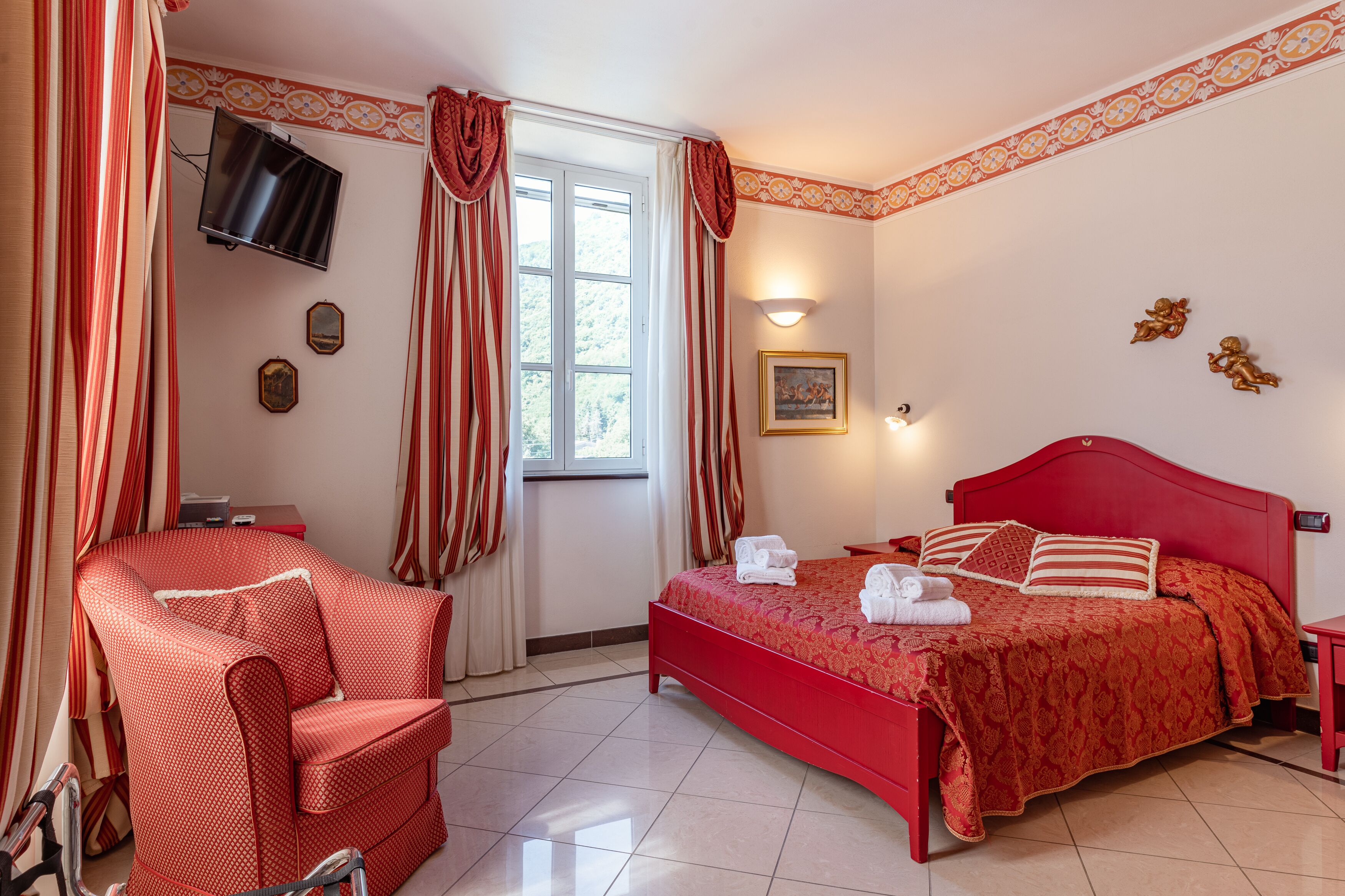 Een hotelkamer met een rode bed, een rode fauteuil, een aan de muur bevestigde televisie en een raam met gordijnen.