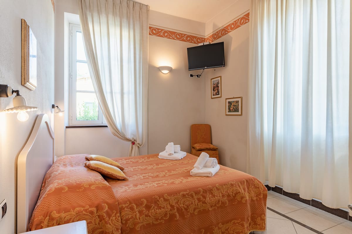 Una habitación de hotel con cama, televisión montada en la pared, ventana con cortinas y una silla.
