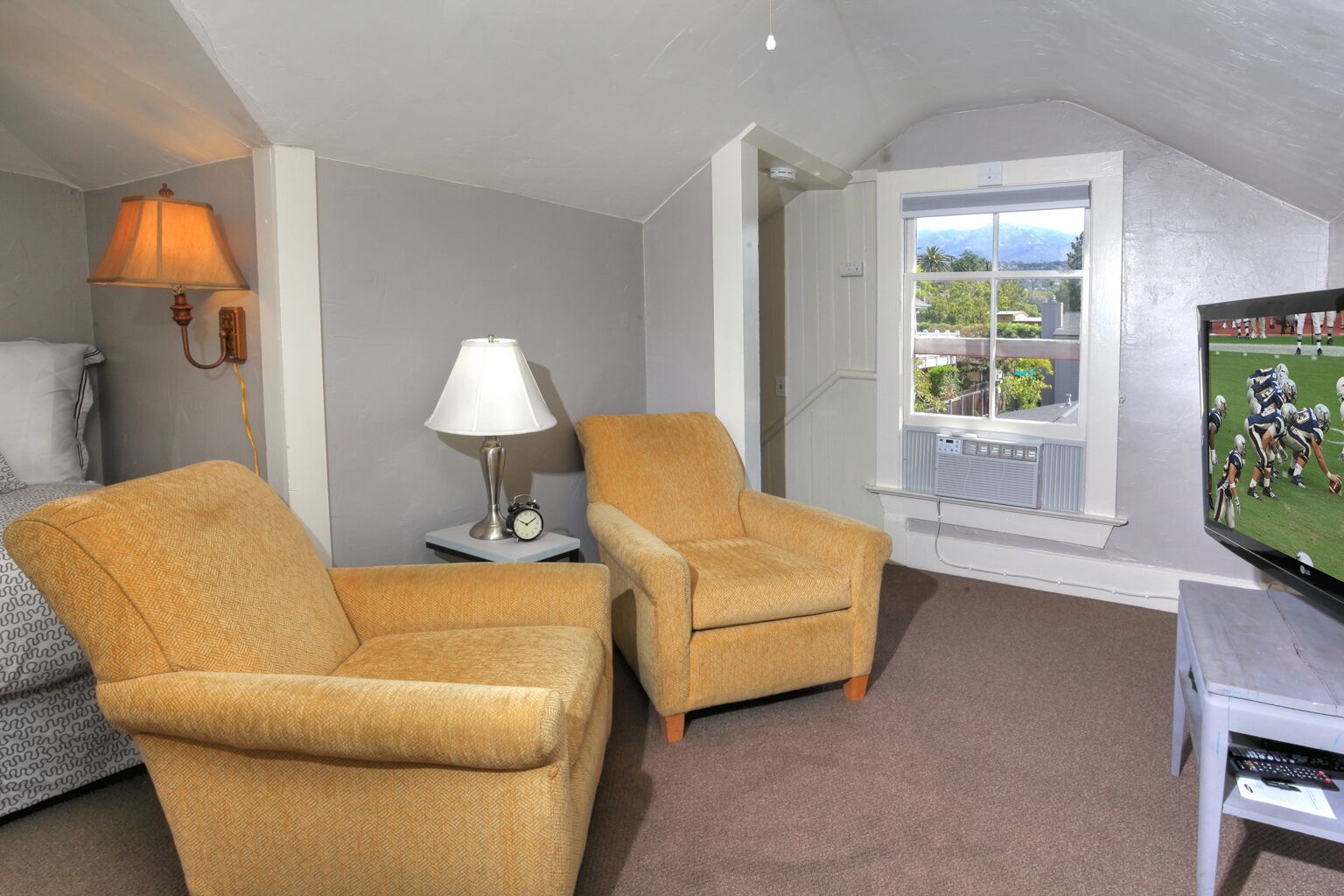 A living room with two yellow armchairs, a table with a lamp and clock, a TV displaying a sports game, and a window with a view of greenery and mountains.