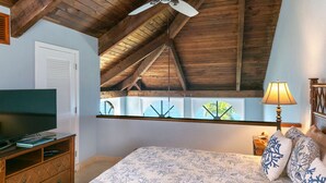 Une chambre avec un plafond en pente, un lit recouvert d’une literie à motifs floraux, un téléviseur posé sur un meuble en bois et une fenêtre donnant sur un paysage verdoyant.