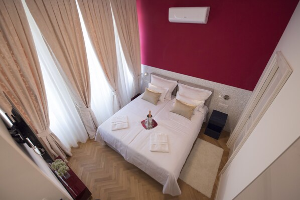 Una cama bien hecha con sábanas y almohadas blancas, una pared roja y una mesita de noche con una botella y vasos.