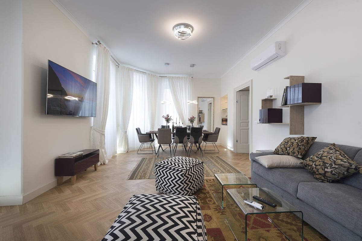 A modern living room with a flat-screen TV, a grey sofa, and a dining area with a table and chairs.