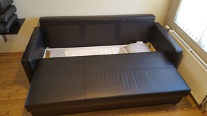 A black sofa bed with a wooden floor and a window with blinds. - Vatnsás 10 (Stykkisholmur)