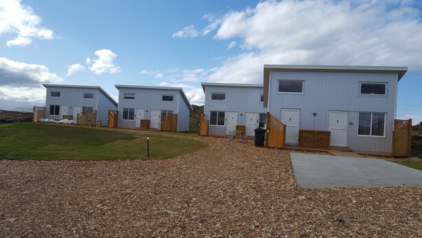 A row of modern, single-story buildings with numbered doors, a gravel driveway, and a grassy area. - Vatnsás 10 (Stykkisholmur)