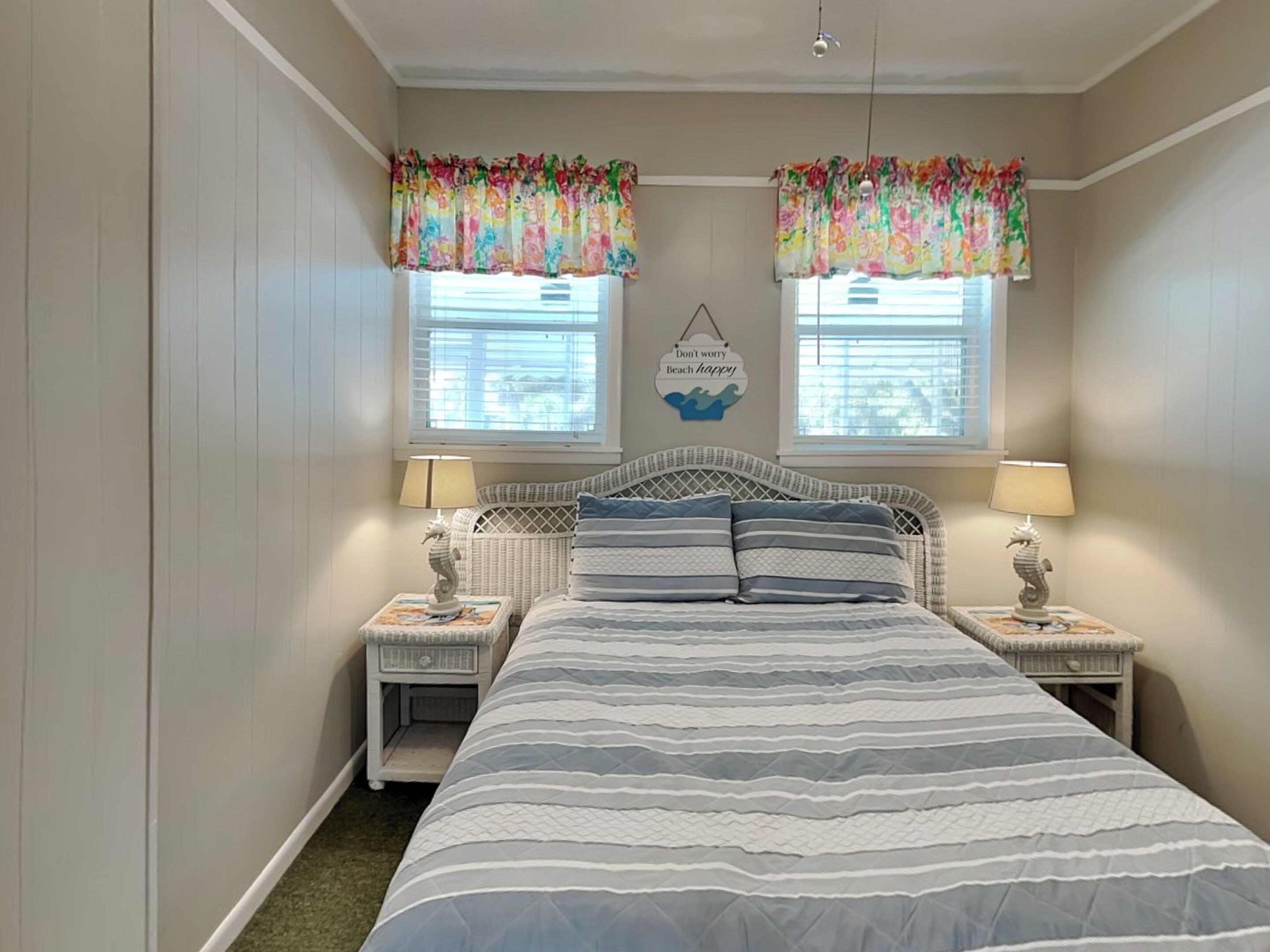 A bedroom with a bed, two nightstands, two lamps, a wall-mounted sign, and two windows with floral curtains.