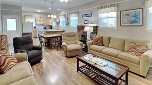 A spacious living room with a sofa, armchairs, and a coffee table. A kitchen area with a dining table and chairs is visible in the background.