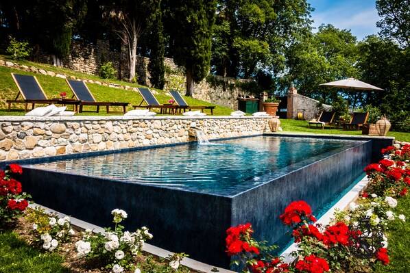A rectangular swimming pool with a stone border, surrounded by white and red flowers, and two lounge chairs with umbrellas.