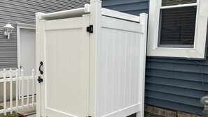 A white outdoor storage unit with a door and a handle, positioned against a house with a window and a white fence.