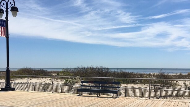 A wooden boardwalk with a bench and a lamp post, overlooking a beach with dunes and a clear sky.