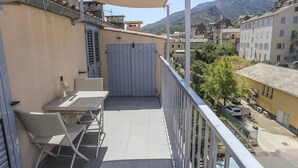 Un balcon avec une table et des chaises, une porte fermée, et une vue sur une rue et des montagnes en arrière-plan.