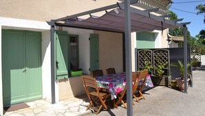 Un espace extérieur abrité, aménagé pour les repas, avec une table et des chaises, une pergola et une maison aux volets verts et au toit en tuiles.