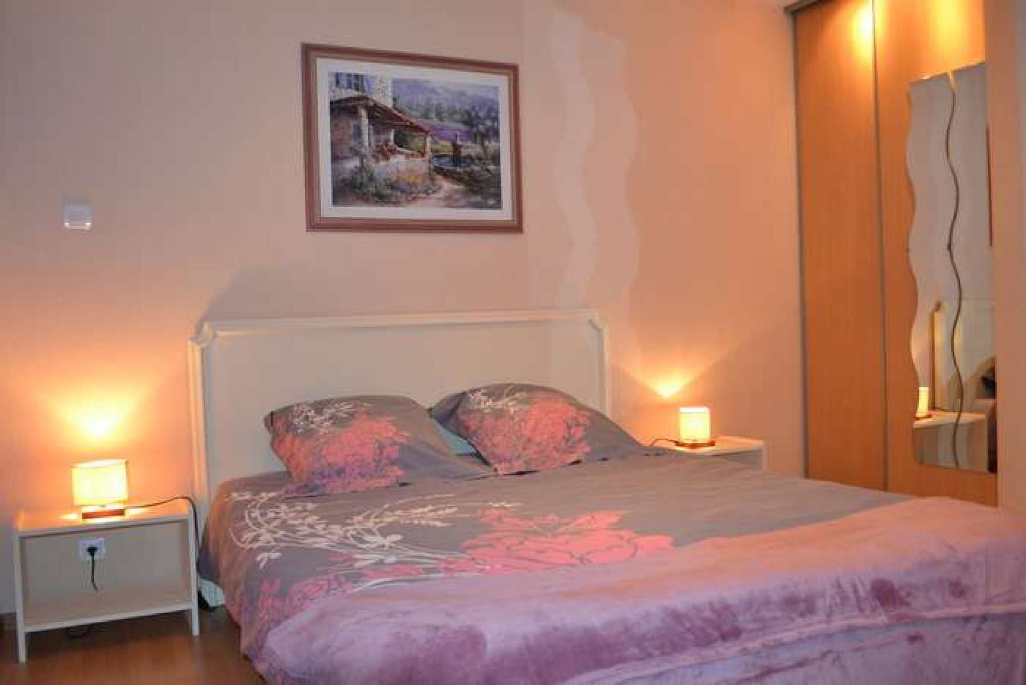 A bedroom with a bed, two bedside lamps, a nightstand, a painting on the wall, and a mirror on the wall.