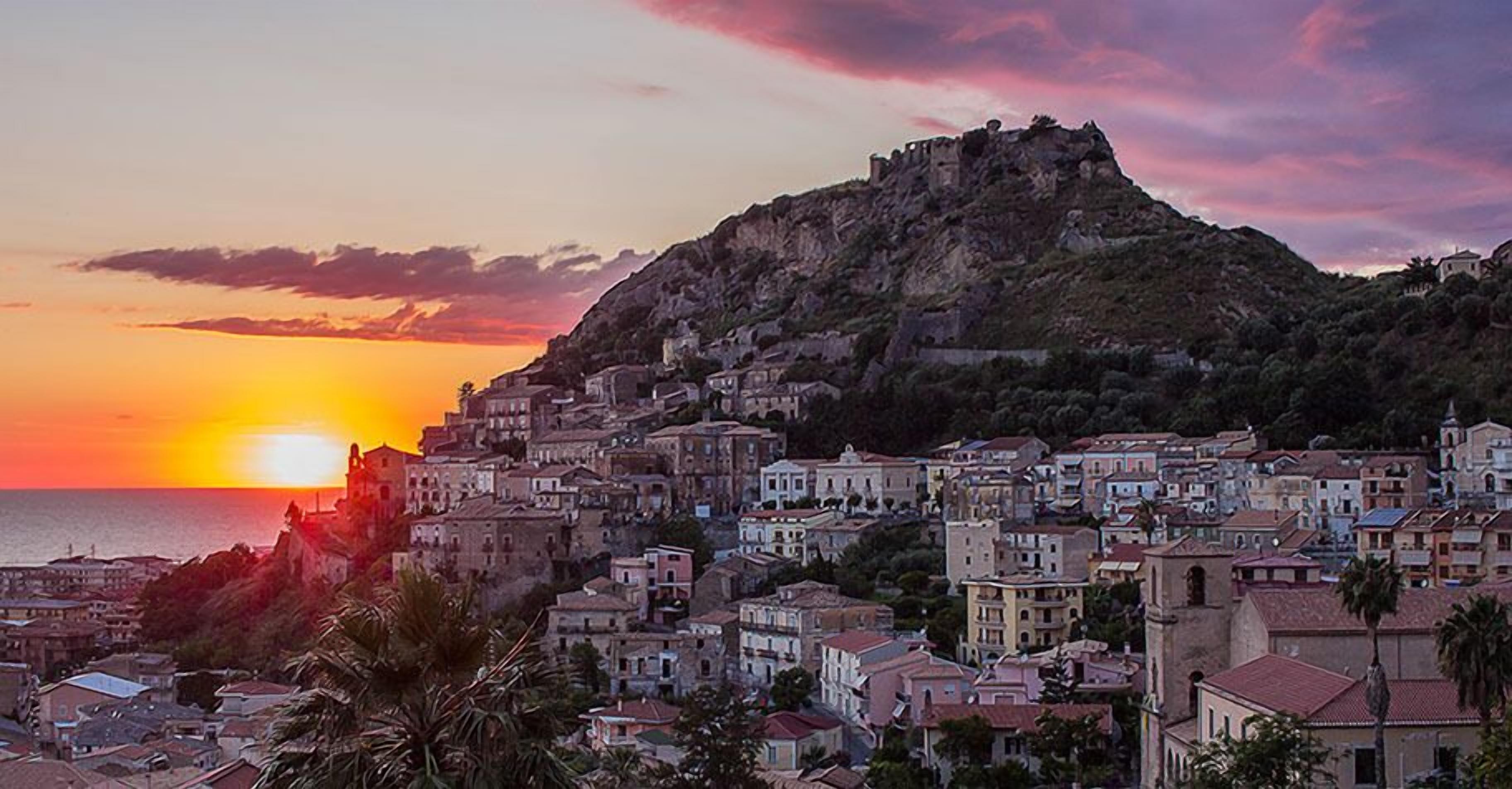 A coastal town at sunset with a prominent hill and a church on top.