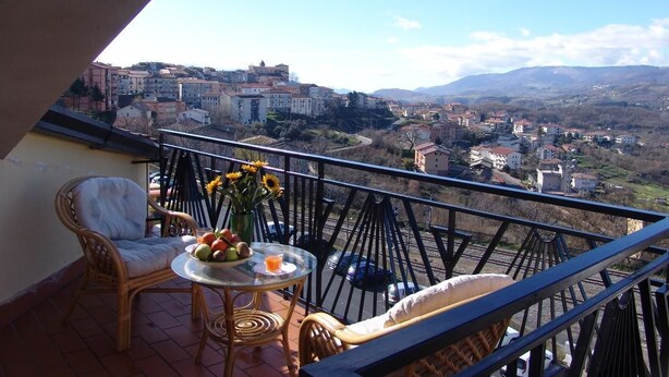 A balcony with wicker chairs, a small round table with a vase of flowers, and a view of a town with buildings and mountains in the distance. - Il Tulipano (Rogliano)
