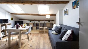 Una sala de estar acogedora con piso de madera, un sofá gris, una mesa de comedor con sillas, una estantería y un tragaluz.
