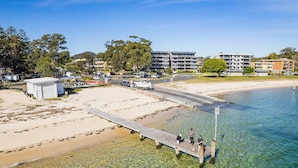 A beachside area with a pier extending into clear water, surrounded by buildings and greenery. - Mistral Court 2, 17 Mistral Close - little beach water views & Wifi (Nelson Bay)