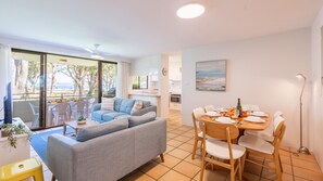 A modern living room with a grey sofa, dining table with chairs, and a view of the beach through large windows.
