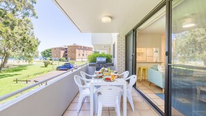 A balcony with a table set for dining, a grill, and a view of a building and park.