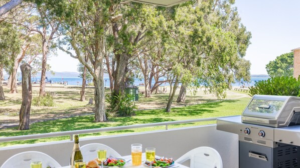An outdoor dining area with a grill, table with food and drinks, and a view of the beach and trees.