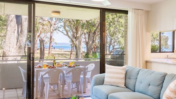 A balcony with a table set for dining, overlooking a beach with trees and the sea. - Mistral Court 2, 17 Mistral Close - little beach water views & Wifi (Nelson Bay)