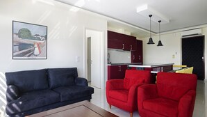 Een woonkamer met een donkerblauwe bank, twee rode fauteuils en een houten salontafel. In de achtergrond is een keuken zichtbaar met donkerrode kastjes en een wit aanrecht.
