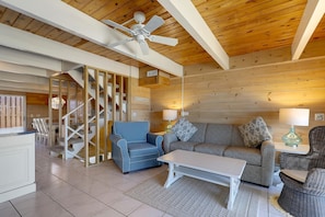 Interior acolhedor de uma cabana com teto de madeira, um ventilador de teto, uma poltrona azul, um sofá cinza e uma cadeira de vime.