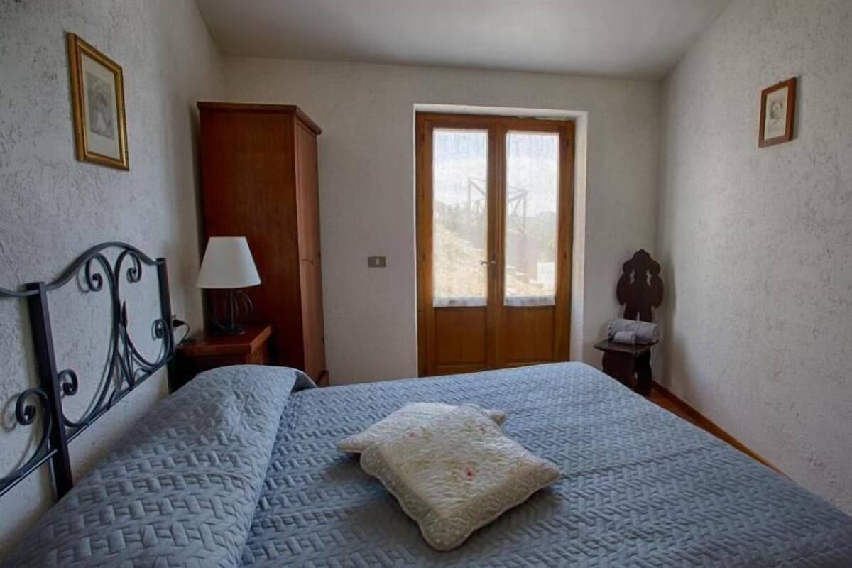 Ein Schlafzimmer mit einem Bett, einem Holzschrank, einem Nachttisch mit Lampe, einer Tür mit Blick und einem kleinen Tisch mit Stuhl.