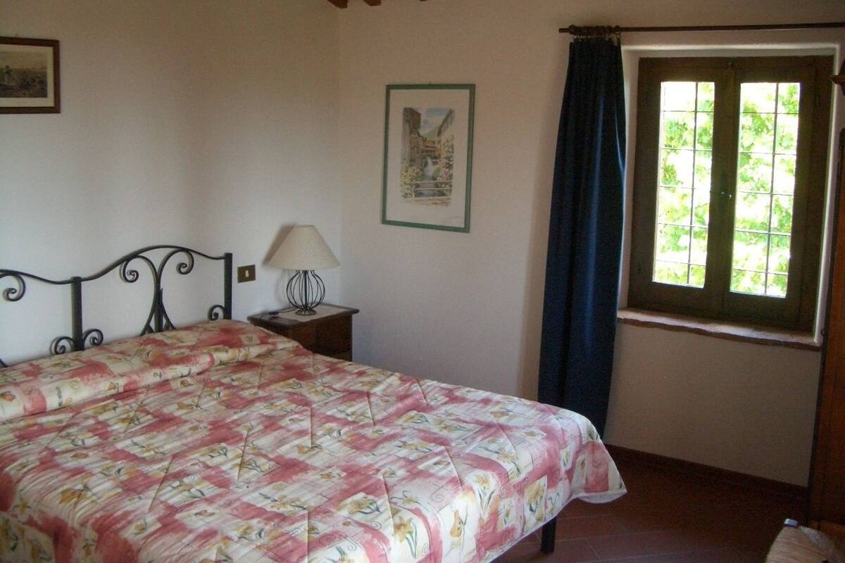 Ein Schlafzimmer mit einem Bett, einem Nachttisch, einer Lampe, einem Fenster mit Vorhängen und einem gerahmten Bild an der Wand.